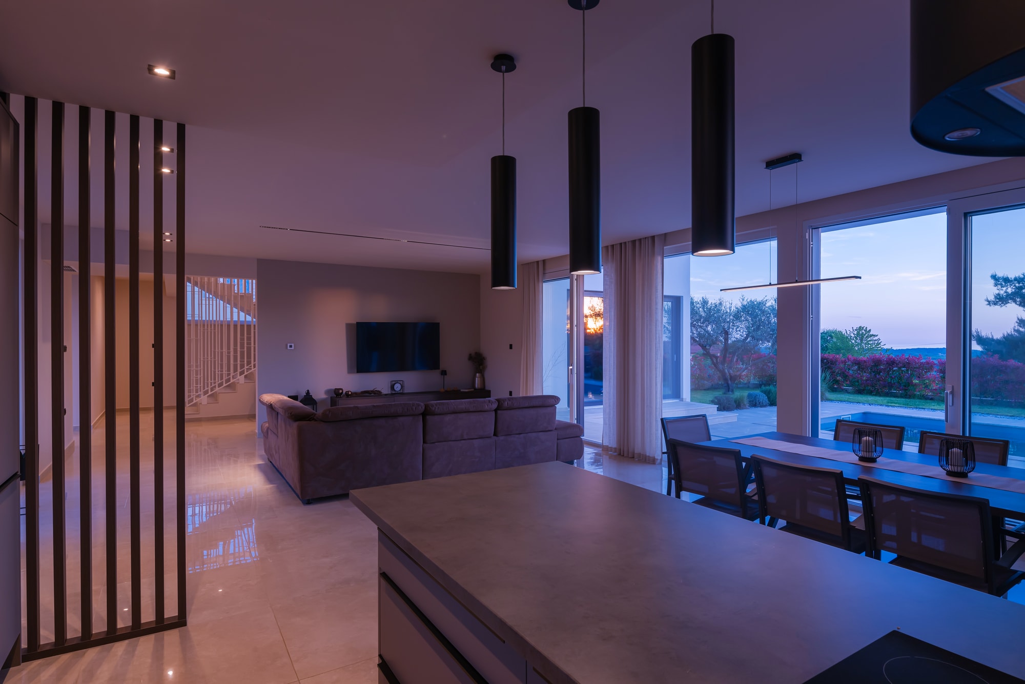 A modern open-plan living and dining space at dusk, a garden view through full-height glass